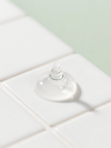Extreme macro close-up of a clear gel skincare droplet on a clean white ceramic tile, bright high-key studio lighting, soft shadows, sharp focus, 50mm