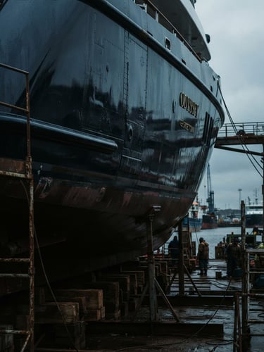 Low-key, dramatic photograph of a superyacht hull in dry dock, showing structural details and a dark, reflective surface, under cold overcast harbor light, 35mm film.