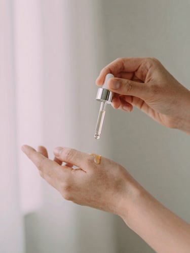 Hands gently applying three drops of botanical oil from a glass dropper bottle onto clean skin, soft morning light, shallow depth of field, muted pink and sage green tones, 35mm.