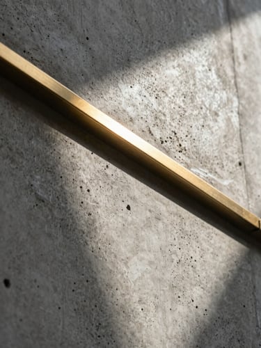 Low-angle architectural detail of raw textured concrete meeting polished brass trim, sharp diagonal shadow casting across the surface under high-contrast sunlight.