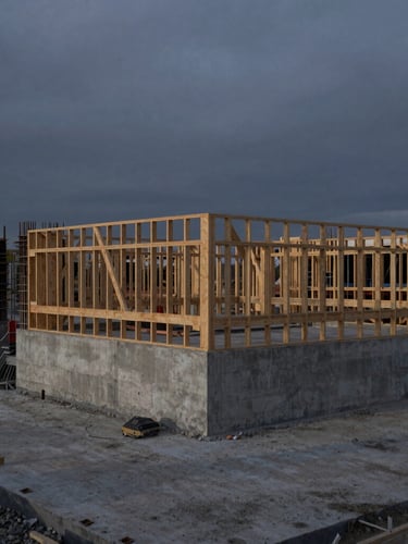 Wide-angle dramatic construction site at dusk, concrete foundation poured, timber framing visible against a deep gray overcast sky, sharp focus, 35mm
