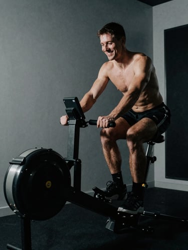 Focused non-smiling athlete training on a premium stationary ergometer in a low-key lit private gym, dramatic side shadows, high contrast, cool tones
