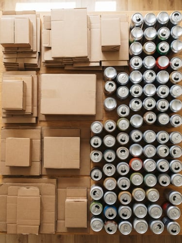 A top-down flatlay of clean, dry cardboard boxes and aluminum cans sorted on a light wooden table, bright natural daylight, 35mm lens.
