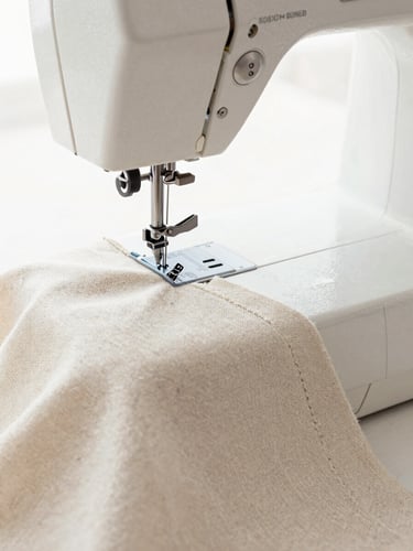 Macro detail of a high-end sewing machine stitching a clean seam on neutral ivory linen fabric, bright natural workshop light, 50mm