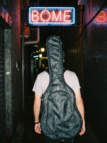 Candid street-style shot of a musician carrying a guitar case down a dark, wet alleyway illuminated by a flickering neon sign.