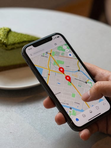 Close-up of a hand holding a smartphone displaying a map with active coral pins, next to a fresh matcha cake on a light gray table, warm natural light.
