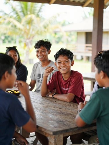Medium shot of active Indonesian village youth collaborating outdoors at a wooden table, warm natural daylight, bright optimistic smiles, 35mm lens style.