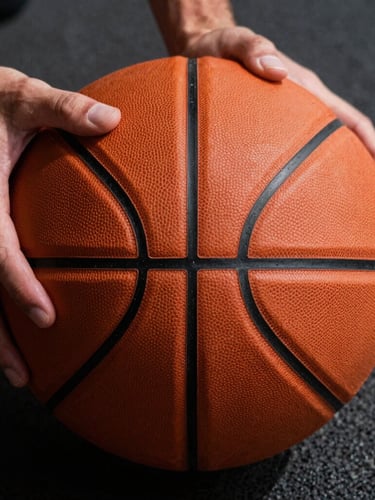 Close-up macro shot of hands gripping a worn orange leather basketball with sharp black seams, dramatic overhead court lighting, dark asphalt background.