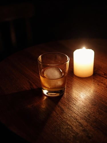 Moody low-key photography of a candle-lit wooden table in a dark cellar tavern, a glass of amber liquid with a single large ice sphere, deep shadows, warm glow.