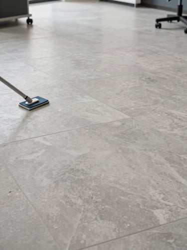 Wide overhead shot of a commercial porcelain tile floor mid-crystallization treatment, applicator pad visible on the surface, diffuse available light in a real office space, neutral gray tile surface with visible texture variation, no people in foreground