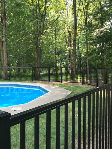 Black aluminum safety fence surrounding an inground swimming pool in a lush wooded backyard.