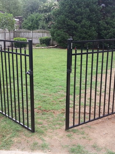 Open black ornamental aluminum fence gate leading to a green grassy backyard with a wooden privacy fence.