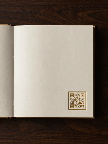 Close-up flat-lay of the back cover of a memorial book, a small square QR code embossed in the lower right corner of an ivory cloth cover, gold foil border around the code catching directional soft light from the upper left — minimal composition, generous breathing space, dark wood surface beneath
