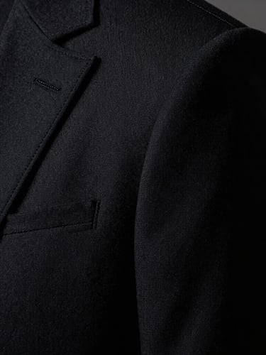 Extreme close-up of a structured shoulder seam and lapel of a premium black wool suit jacket, highlighting the immaculate stitching and heavy fabric texture under harsh, cinematic natural light.