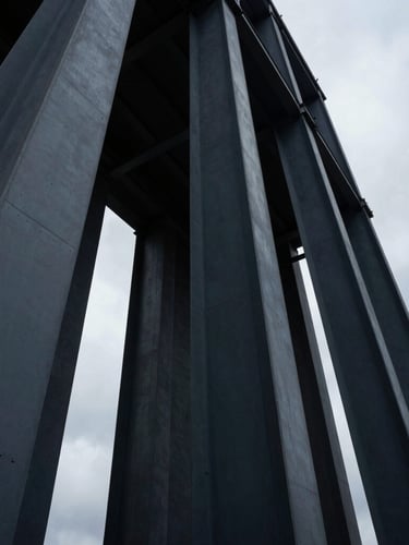 Low-angle architectural photograph of massive structural steel columns rising into a dramatic overcast sky, deep slate tones