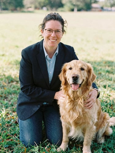 Candid warm editorial portrait of a professional specialized educator and a gentle golden retriever sitting together in a sunlit outdoor meadow, 35mm film look