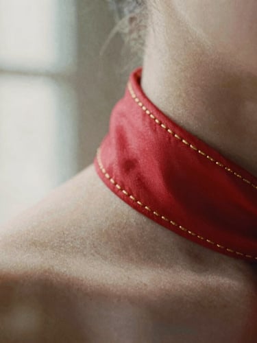 Extreme close-up of delicate gold hand-stitching on a scarlet silk collar, soft directional natural light with deep soft shadows, 35mm film grain