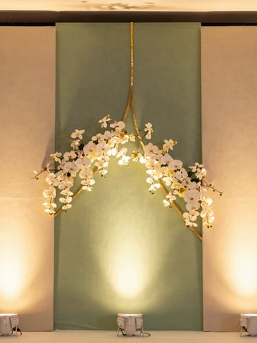 A tall portrait shot of a contemporary Indian wedding stage backdrop, featuring asymmetric sage green panels, delicate gold-leaf trim, and cascading white orchids under soft warm spotlighting