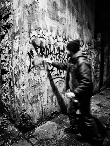 Raw flash photography, high contrast black and white, gritty urban environment, direct harsh light, 28mm wide-angle lens, showing a blurred figure spray-painting a defiant message on a concrete wall.