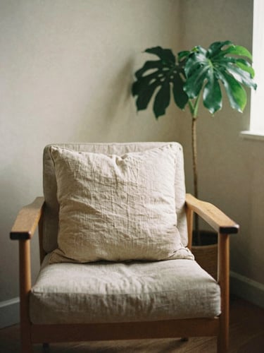 Softly lit empty therapy room with a wooden chair, linen pillows, and a lush green plant in the corner, warm diffused morning light, muted earth tones