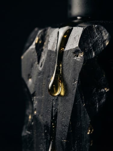 Macro photograph of a thick, golden droplet of perfume oil slowly sliding down the hand-carved obsidian glass bottle, dramatic chiaroscuro lighting, deep black background with sharp gold highlights.