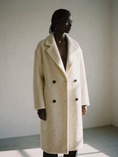 Soft focus silhouette of a model in a heavy cream cashmere coat, standing in an architectural corner of an empty studio, diffused daylight, warm shadows