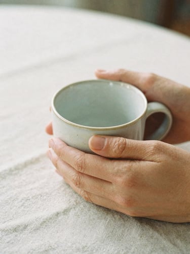 Close-up of hands holding a warm ceramic mug on a textured linen tablecloth, soft morning daylight, muted cream and clay tones, shallow depth of field, 35mm film