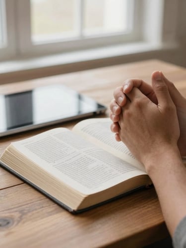 Hands folded in prayer over an open Bible and a digital tablet on a wooden table, soft window light