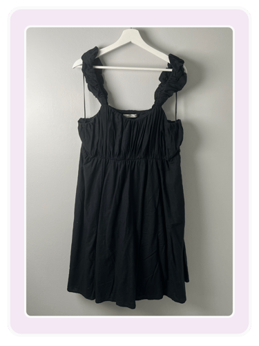 Black sleeveless mini dress with ruffled straps and a gathered bodice hanging on a white hanger.