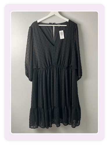 Black long-sleeve Swiss dot mini dress with a V-neckline hanging on a white hanger against a gray wall.