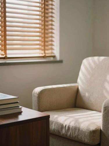 A minimalist, warm clinical consultation room with soft sunlight filtering through wooden blinds, a comfortable beige armchair, a stack of books on a dark wood table, shallow depth of field, 35mm photography