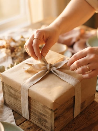 Close-up of hands carefully tying a linen ribbon around a beautifully wrapped wooden gift box, warm golden-hour window light, shallow depth of field