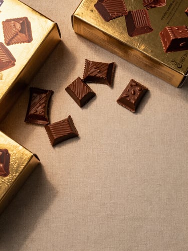 Macro close-up overhead flat-lay of premium packaged confectionery boxes and unwrapped chocolate pieces on textured linen, warm tungsten studio light from upper left casting soft shadows, gold foil labels visible, arranged off-center with generous negative space at bottom right