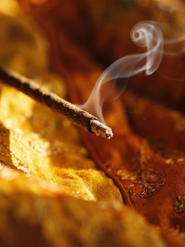 luxurious editorial photography of a single hand-rolled incense stick, delicate curl of pale grey smoke rising in dramatic golden-hour side light, rich saffron background, macro depth of field, 35mm