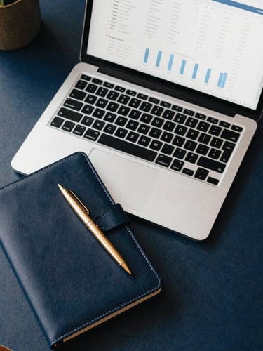 Overhead flatlay of a modern designer workspace, sleek laptop showing clean analytics charts, premium leather notebook with a gold pen, soft daylight, deep navy and warm gold tones, editorial workspace photography