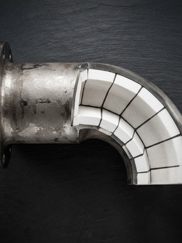 Sharp orthographic studio shot of a heavy steel pneumatic conveying elbow, cutaway showing internal white ceramic tile lining, high-contrast spotlighting on a dark slate background, macro depth of field.
