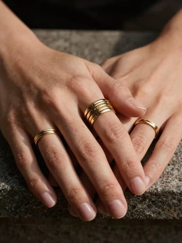 Cropped environmental shot of hands wearing multiple stacked 18k gold rings resting on a textured stone plinth, warm directional light