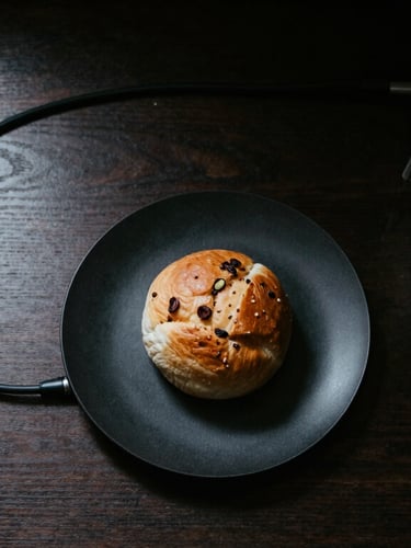 Moody top-down shot of a minimalist workspace, a dark wood desk, clean cable routing, and a matte black plate with a fresh cardamom bun next to an espresso machine portafilter, high contrast.