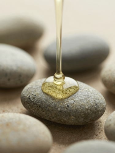 Close-up macro shot of warm herbal oil dripping slowly onto smooth basalt stones, soft diffused morning light, muted sage and sand tones, shallow depth of field