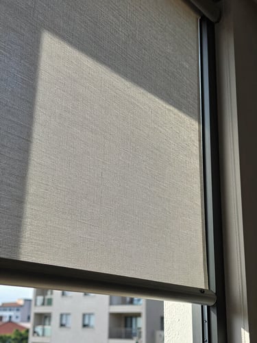 Extreme close-up of precision-cut roller blind fabric hanging against a tall residential window in Mexico City, cool north-facing daylight raking across the woven texture, aluminum bottom rail perfectly level, architectural plaster reveal visible at edge