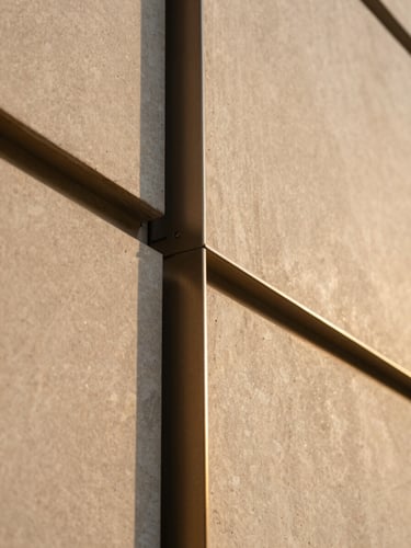 Close-up detail of a contemporary building facade — precision-cut stone cladding panels meeting a bronze anodized aluminum frame, shallow depth of field isolating the material transition, warm golden hour raking light revealing surface texture and shadow depth, architectural material study framing