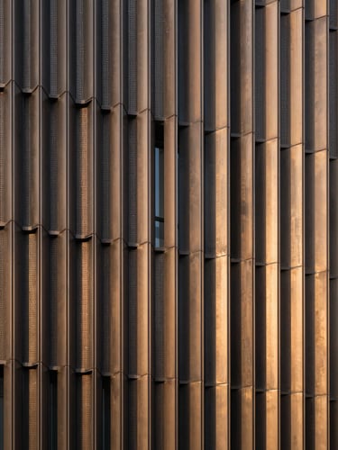 Close-up architectural photograph of a completed contemporary building facade, dark aluminum fins and perforated bronze panels in precise geometric rhythm, controlled studio-quality light revealing material texture
