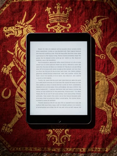 Top-down flatlay of a modern tablet displaying an e-book, resting on a rustic medieval stone surface with subtle red and gold heraldic linen textures, dramatic high-contrast side lighting, 35mm