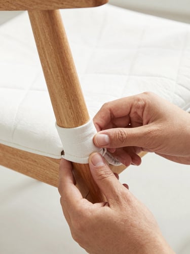 Crisp editorial close-up of hands carefully wrapping a wooden chair leg with thick quilted padding, bright natural daylight, sharp focus