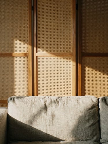 Architectural close-up of a warm wood and rattan folding screen dividing a sunlit workspace, soft afternoon light casting textured shadows on a linen sofa, muted earth tones, 35mm