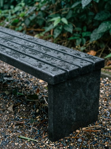 Close-up of a freestanding storage bench made of dense dark resin, wet from morning dew, placed on a gravel path, 35mm