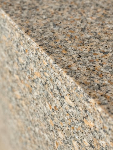 Extreme close-up overhead of a polished granite slab edge profile, directional daylight raking across the surface to reveal crystal grain and finish texture, warm neutral tones, no color correction, full-frame crop showing material reality