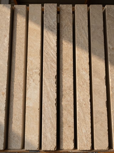 Overhead flat-lay view looking down a row of upright stone slabs in a warehouse rack, warm directional daylight raking across the faces to reveal surface variation and veining differences between adjacent slabs, warm neutral tones, no color correction