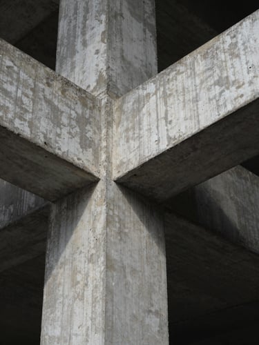 Fotografia em perspectiva de um ponto de uma viga de concreto aparente cruzando um pilar de aço escovado, luz natural difusa lateral, sombras geométricas marcadas.