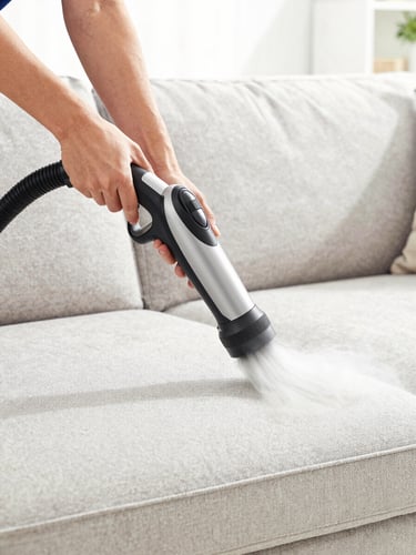 A close-up of a technician's hands using a professional steam-cleaning wand on a fabric sofa, lifting dirt, high-key natural daylight.